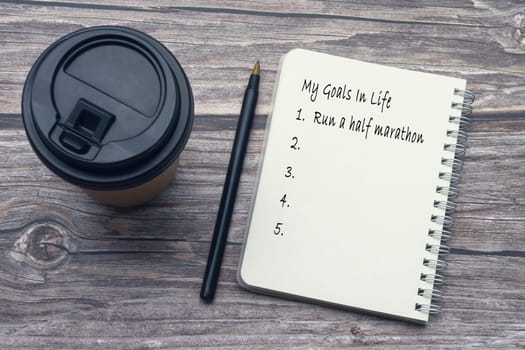 My goals in life list on notepad with pen and disposable coffee cup on wooden desk.