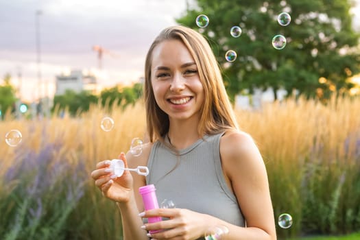 A joyous young woman with long, flowing hair creates soap bubbles at sunset in the park.