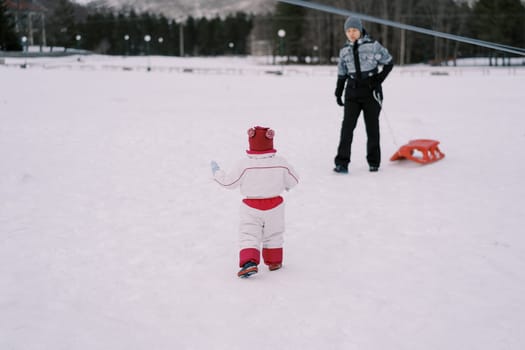 Little girl goes to her mother standing with a sled in the snow. Back view. High quality photo