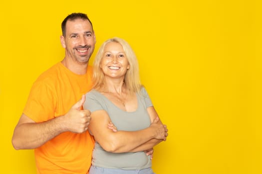 Happy adorable Caucasian couple in their 40s hugging and raising thumbs up in sign of approval and trust, isolated on yellow studio background