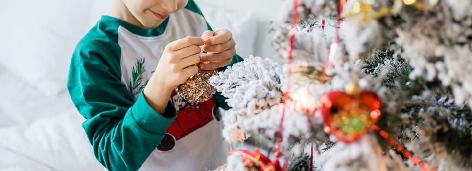 Child decorates Christmas tree. The boy is holding a Christmas toy. Morning before christmas at home. Generation alpha and gen alpha children