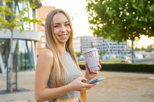 Cheerful young woman smiles at camera holding a cup and smart phone in her hands, copy space