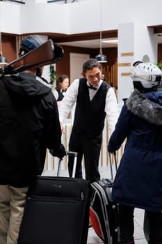 Couple in winter clothing arriving in hotel reception and young asian bellboy assists them with their luggage. Male employee handling suitcases of visitors at arrival in ski mountain resort.