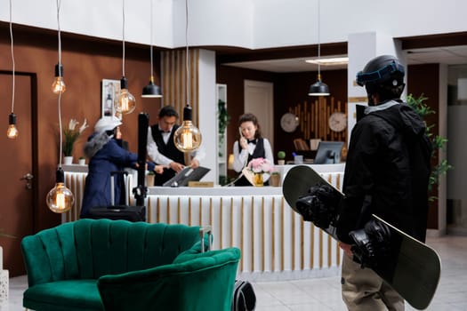 Receptionist provides service to female customer while man with winter clothing enters ski hotel reception. Young travelers with skiing and snowboarding gear arrive in lounge area for check-in.
