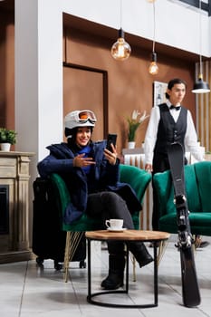Asian woman in hotel lobby holding smartphone while wearing ski goggles and winter gear. Female customer grasping digital phone device seated comfortably on sofa in luxury ski mountain resort.