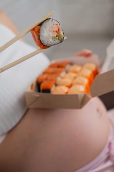 A pregnant woman sits on the sofa and eats rolls from a box. Food delivery. Close up of belly