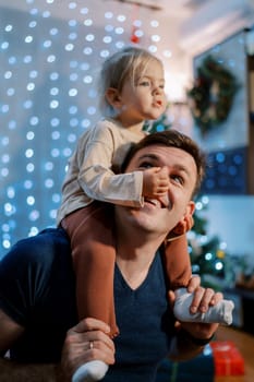 Little girl touching dad nose while sitting on his shoulders near the decorated Christmas tree. High quality photo