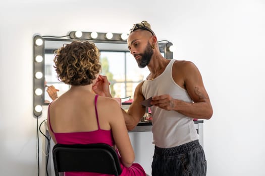 Male makeup artist apply lipstick for woman who in front of mirror. Bearded gay professional make-up artist on backstage at work. MUA for female client.