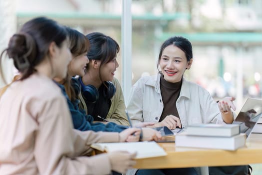 Group of young Asian college students sitting on a bench in a campus relaxation area, talking, sharing ideas, doing homework or tutoring for the exam together.