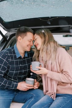 Smiling boyfriend and girlfriend touch foreheads while sitting with coffee mugs in car trunk. High quality photo