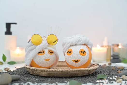 Two orange fruits in face mask and towel on black pebbles. Spa treatment and self care concept.