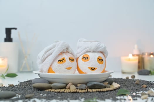 Two orange fruits in face mask and towel on black pebbles. Spa treatment and self care concept.