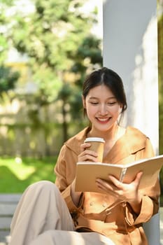 Relaxed young Asian woman drinking coffee and reading book at outdoor.