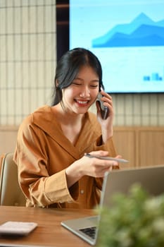 Cheerful Asian female employee looking at laptop screen and talking on mobil phone.