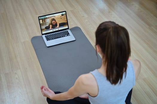 Back view of young woman sitting on yoga mat watching active sports workout video lesson on laptop.