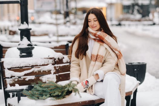 A girl with long hair in winter sits on a bench outside with a bouquet of fresh fir branches.