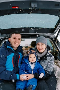 Laughing mom and dad with little girl sitting in car trunk in winter forest. High quality photo