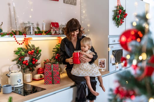 Christmas kitchen mom daughter. They are sitting on a table and drinks from a red mug in a decorated kitchen for christmas