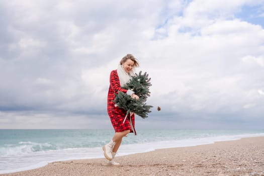 Blond woman holding Christmas tree by the sea. Christmas portrait of a happy woman walking along the beach and holding a Christmas tree in her hands. Dressed in a red coat, white dress