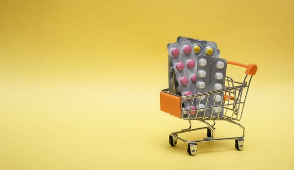 Many medicine pills, tablets in a small supermarket shopping cart on yellow background. concept shopping medicine. Pharmacy theme, capsular tablets with medicinal vitamins
