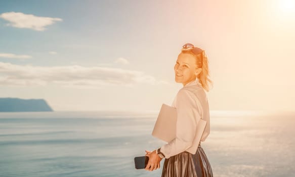 Business woman on nature in white shirt and black skirt. She works with an iPad in the open air with a beautiful view of the sea. The concept of remote work