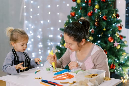Little girl helps her mother apply colorful icing on Christmas cookies. High quality photo