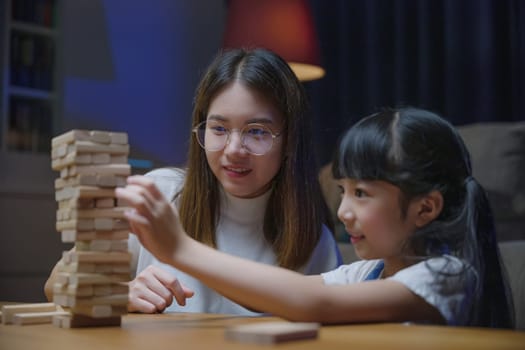 Asian young mother playing game in wood block with little daughter in home living room at night, Smiling woman help teach preschooler kid play build constructor tower of wooden blocks, family funny