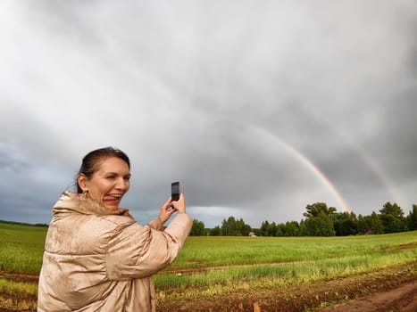 An adult girl in a field and with a stormy sky with clouds takes pictures of a rainbow and takes selfie in the rain. A woman having fun outdoors on rural and rustic nature