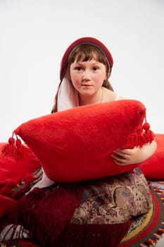 Portrait of Little girl in a stylized Tatar national costume having rest with a red pillow on a white background in the studio. Photo shoot of funny young teenager who is not a professional model