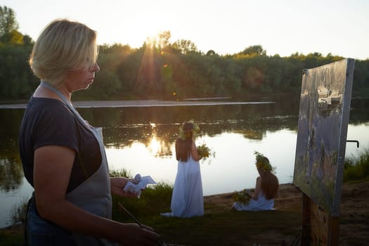 Adult female artist painting picture near water of river or lake in nature and girls in white sundress and flower wreath. Artist and models posing in holiday of Ivan Kupala in nature at sunset