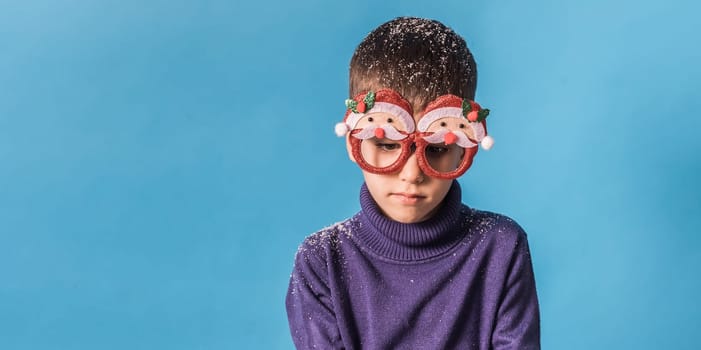 Banner Portrait of sad frustrated boy wearing funny festive santa eyeglasses covering snow eve christmas time isolated over bright blue color background. Generation alpha and gen alpha children