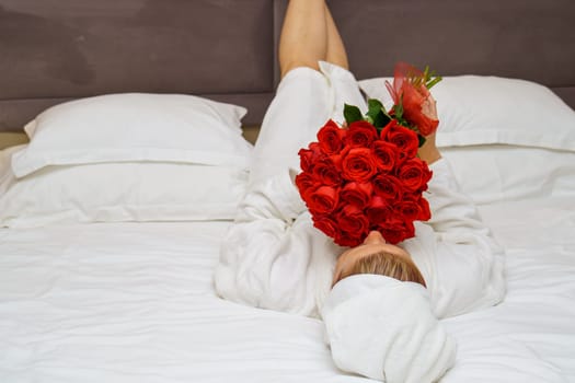 A stunning young woman lying on a luxurious hotel bed, surrounded by a large bouquet of vibrant red roses. She exudes elegance and tranquility, creating a romantic and relaxing ambiance.