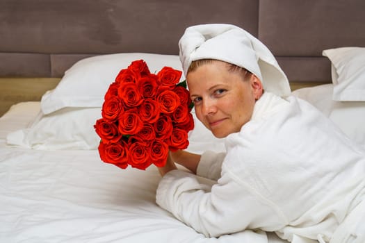 A beautiful young woman in an elegant dress sits on a luxurious hotel bed, holding a large bouquet of red roses, creating a scene of romance and relaxation.