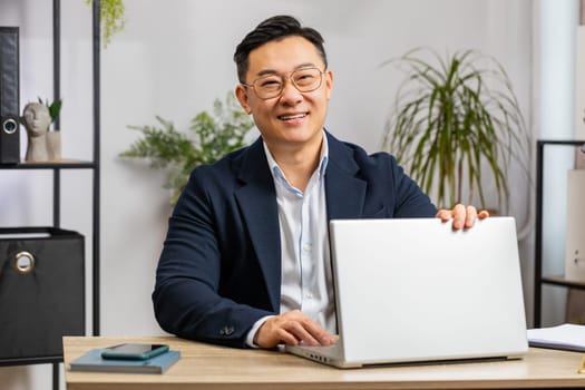 Portrait of happy Asian businessman freelancer at home office modern workplace working on laptop computer. Manager Chinese man in formal suit work on notebook send messages makes online purchases