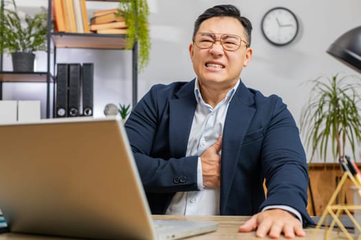 Stressed Asian mature businessman freelancer suffers from tension holding chest with heart pain attack risk of stroke symptom while working at home office workplace desk. Male man in formal suit