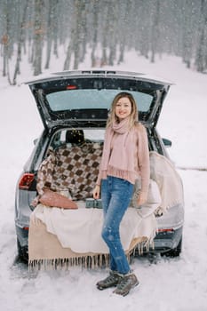 Smiling young woman standing near open trunk of car with blankets and pillows under snowfall in forest. High quality photo
