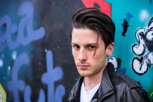 A man in a leather jacket standing in front of a graffiti covered wall