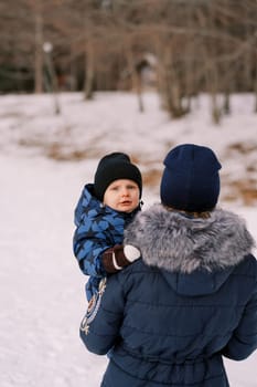 Mother with a little boy in her arms walks along the snow-covered edge of the forest. Back view. High quality photo