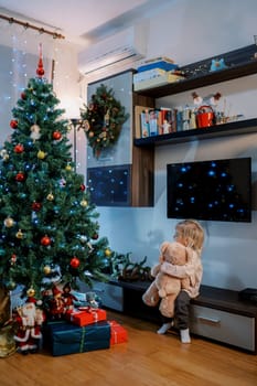 Little girl sits on a bedside table hugging a teddy bear and looks at the Christmas tree. High quality photo