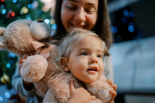 Mom put a teddy bear on the neck of a little girl near the Christmas tree. High quality photo