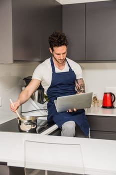 A man sitting on a kitchen counter using a laptop, reading a recipe while stirring a pot. A Handsome, Muscular Man Sitting on a Kitchen Counter with a Laptop