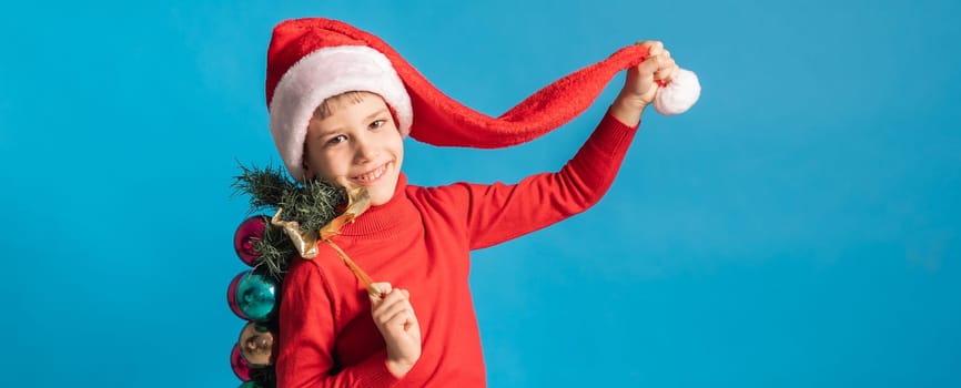 Cheerful child in santa hat hold christmas decorative balls on blue background, Xmas and holidays concept mockup. Generation alpha and gen alpha children