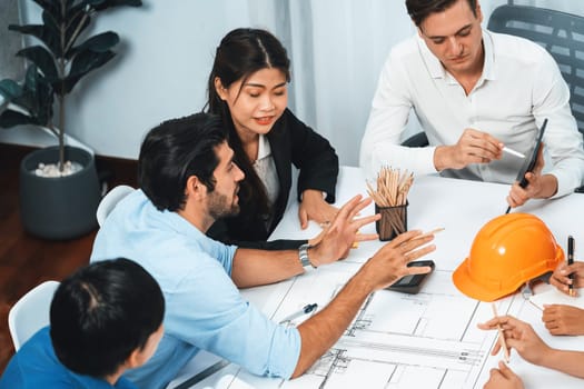 Diverse group of civil engineer and client working together on architectural project, reviewing construction plan and building blueprint at meeting table. Prudent