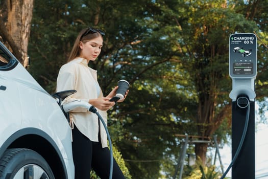 Young woman with coffee cup and sustainable urban commute with EV electric car recharging at outdoor cafe in springtime garden, green city sustainability and environmental friendly EV car. Expedient