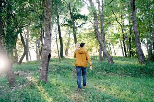 Dad with a little girl in his arms walks through the sunny forest. Back view. High quality photo