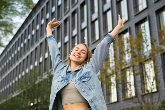 Satisfied young woman with long loose hair widely smiles raising arms upward. Happy lady stretches after work against office building