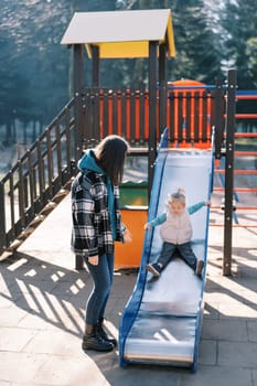 Little girl goes down the slide on the playground next to her standing mom. High quality photo