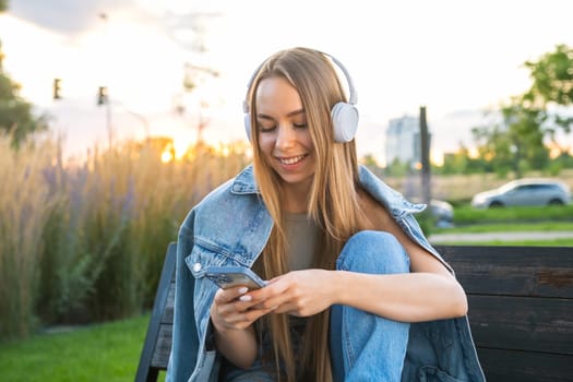 Female student with long blonde hair uses smart phone and listen to music after study sitting on the bench at sunset.