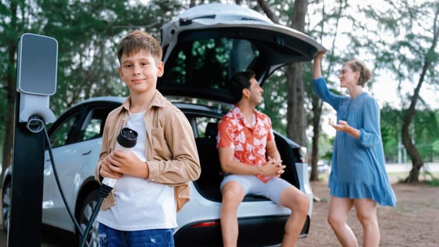 Little boy holding EV charger and point at camera with his family sitting on the trunk in background. Road trip travel with alternative energy charging station for eco-friendly car concept. Perpetual