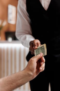 Detailed view of caucasian senior man giving cash to bellhop in exclusive hotel reception for assisting with luggage. Customers providing money tip to bellboy in resort lounge area. Close-up shot.